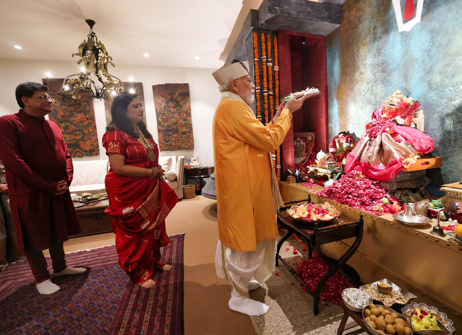 एक व्यक्ति नारंगी रंग के कपड़े पहने हुए, एक मूर्ति के सामने फूल चढ़ा रहा है, जबकि दो अन्य लोग देख रहे हैं।